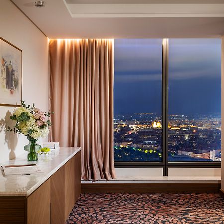 Grand Hotel Millennium Sofia Ngoại thất bức ảnh The photo depicts a modern hotel room or a lounge area with a stunning view of a cityscape at night. On the left, there's a wooden console table with a vase of flowers and a bottle of water. A piece of art is framed on the wall above the table. The r