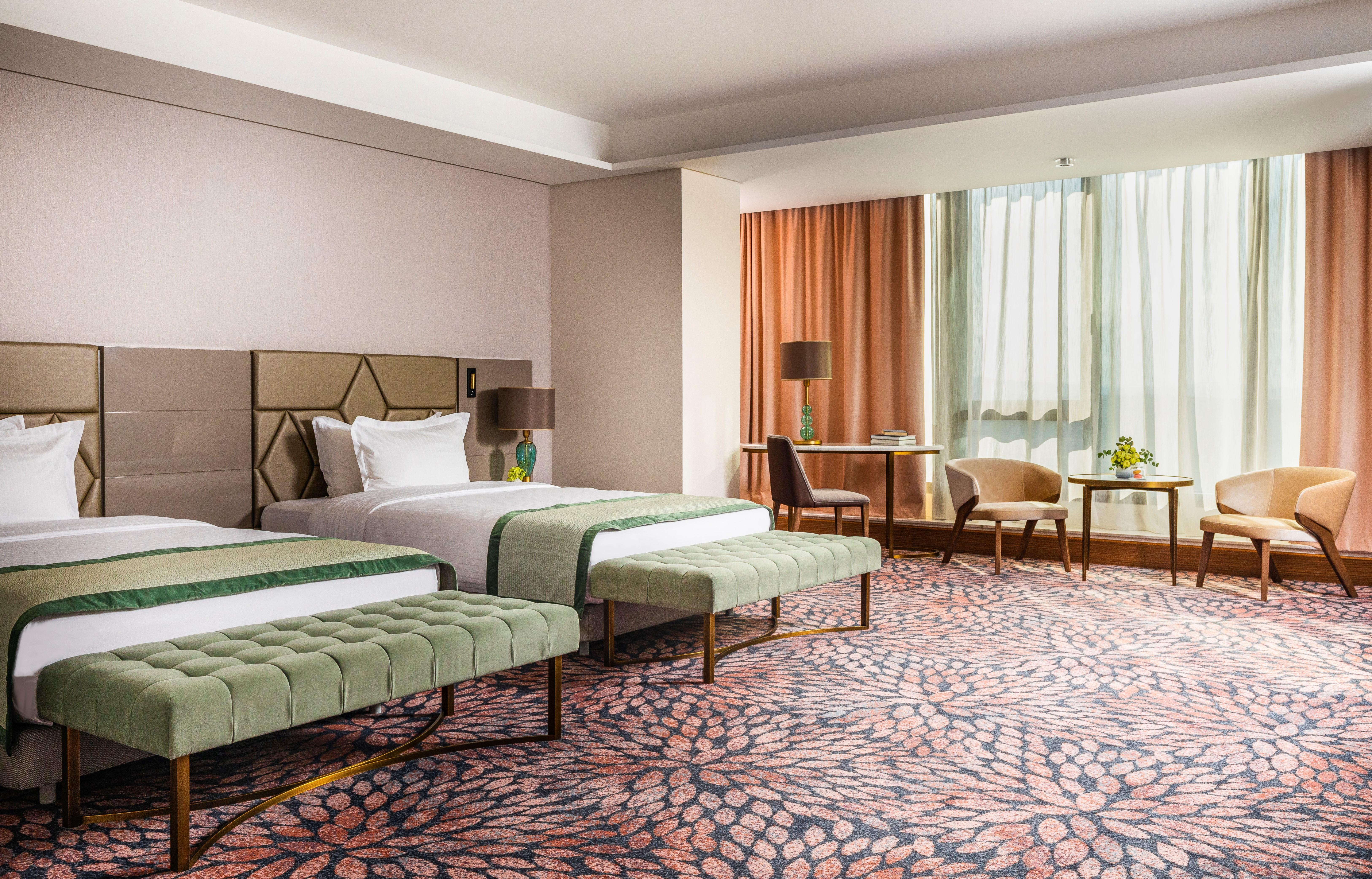 Grand Hotel Millennium Sofia Ngoại thất bức ảnh The image shows a well-decorated hotel room featuring two beds with green striped bedspreads. There is a plush green bench at the foot of each bed. The room has large windows with sheer curtains allowing natural light to enter. There is a cozy seatin
