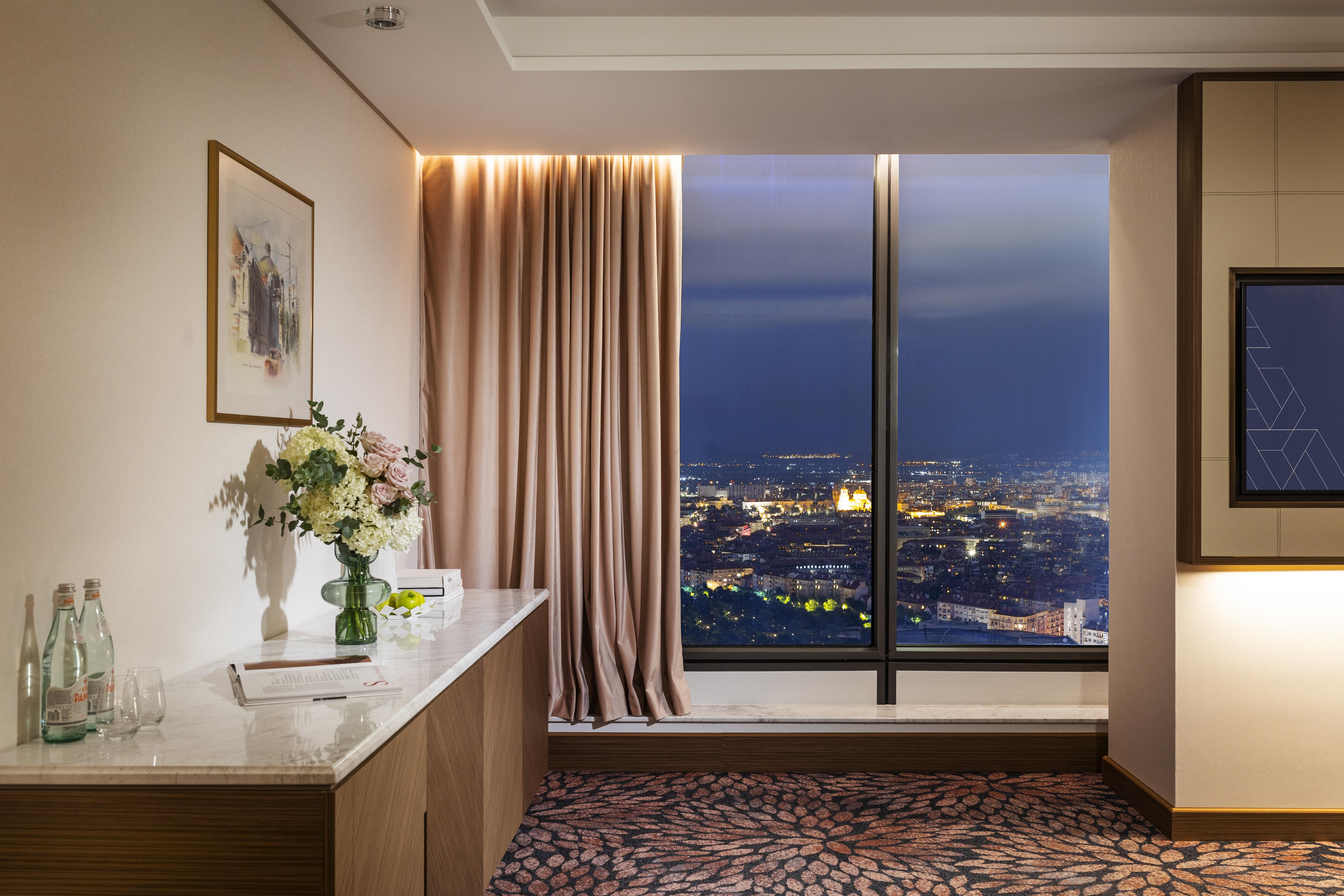 Grand Hotel Millennium Sofia Ngoại thất bức ảnh The photo depicts a modern hotel room or a lounge area with a stunning view of a cityscape at night. On the left, there's a wooden console table with a vase of flowers and a bottle of water. A piece of art is framed on the wall above the table. The r