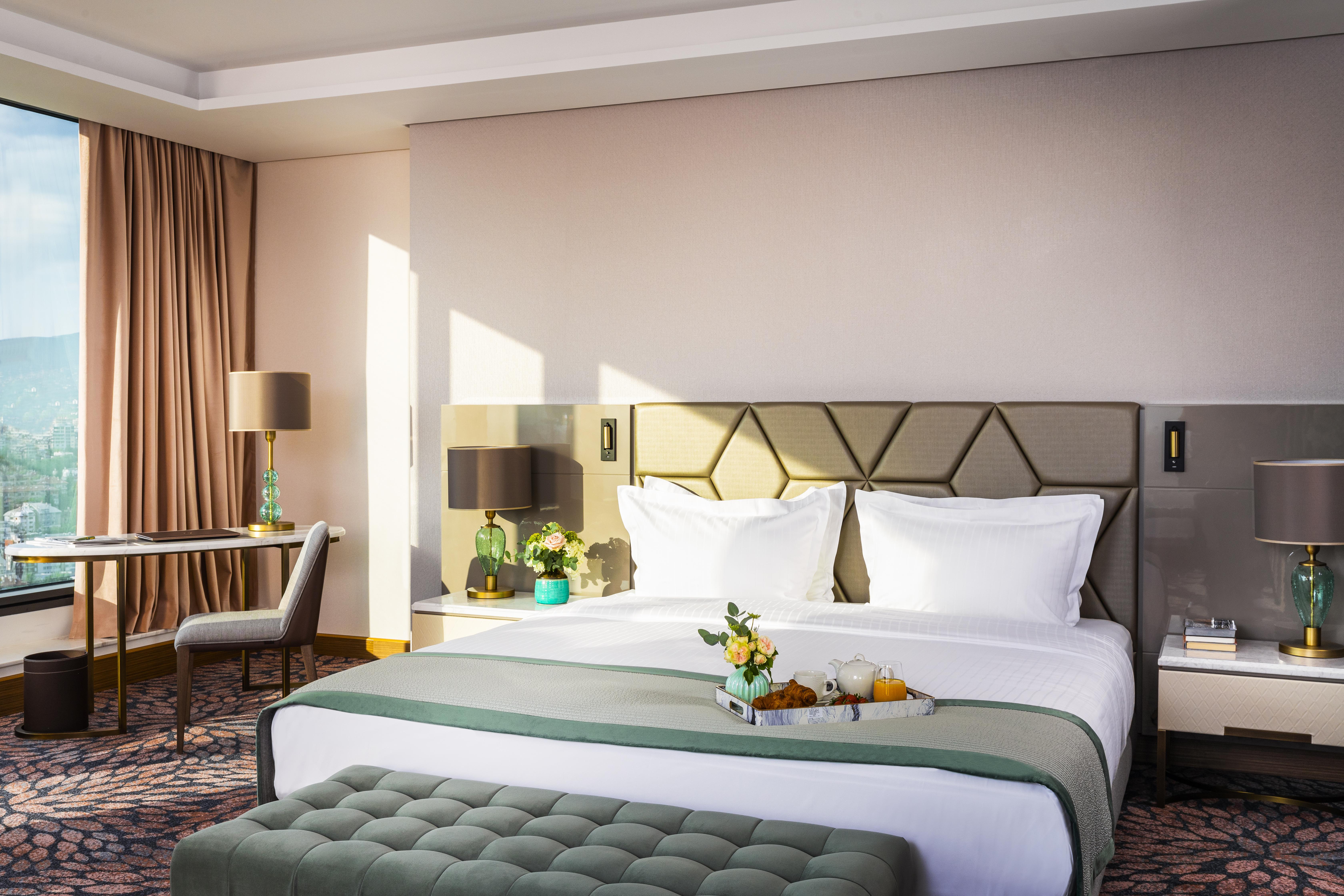 Grand Hotel Millennium Sofia Ngoại thất bức ảnh The photo depicts a stylish hotel room featuring a large bed with white linens and a green decorative throw. Behind the bed is a geometric-patterned headboard. To the side, there are modern bedside lamps and a small table with a vase of flowers. A se