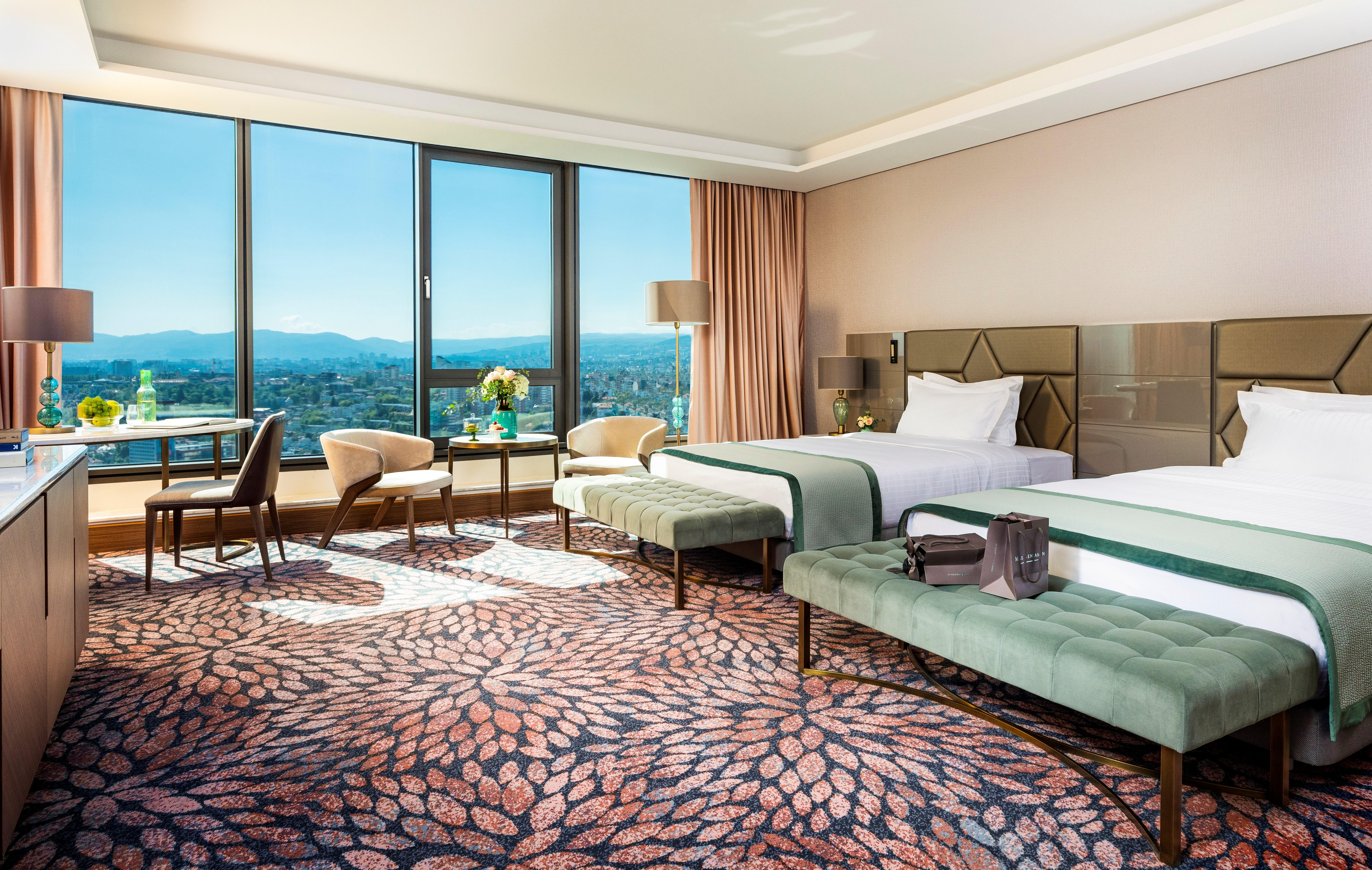 Grand Hotel Millennium Sofia Ngoại thất bức ảnh The photo depicts a luxurious hotel room featuring two beds with elegant bedding. The room has large windows that offer a panoramic view of the city and mountains in the background. There is a decorative area rug with a floral pattern covering the fl