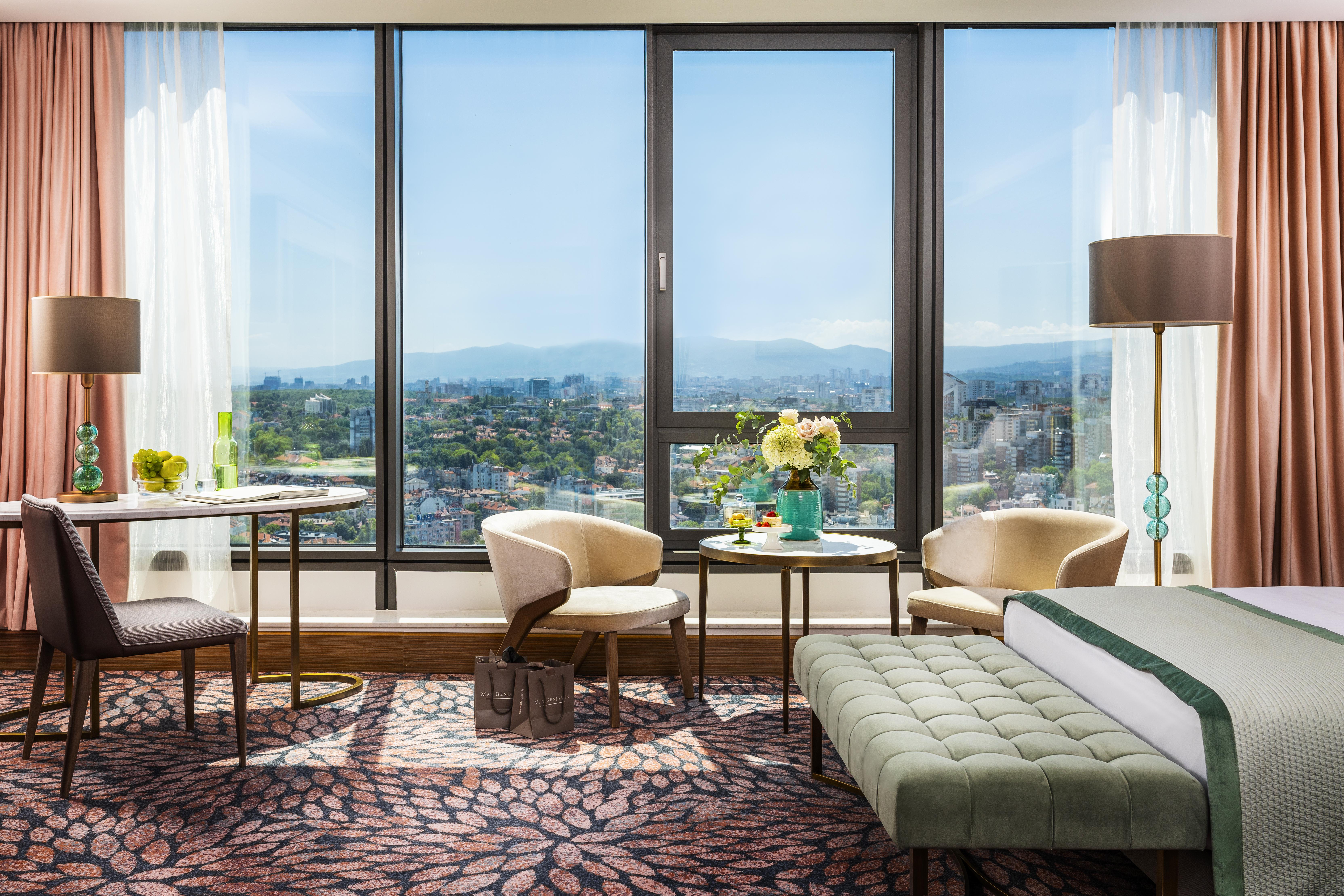 Grand Hotel Millennium Sofia Ngoại thất bức ảnh The photo shows a modern and well-designed hotel room featuring a large window that offers a view of the outdoors, including mountains in the background. Inside the room, there is a neatly made bed with a green accent blanket, a stylish rug with a fl