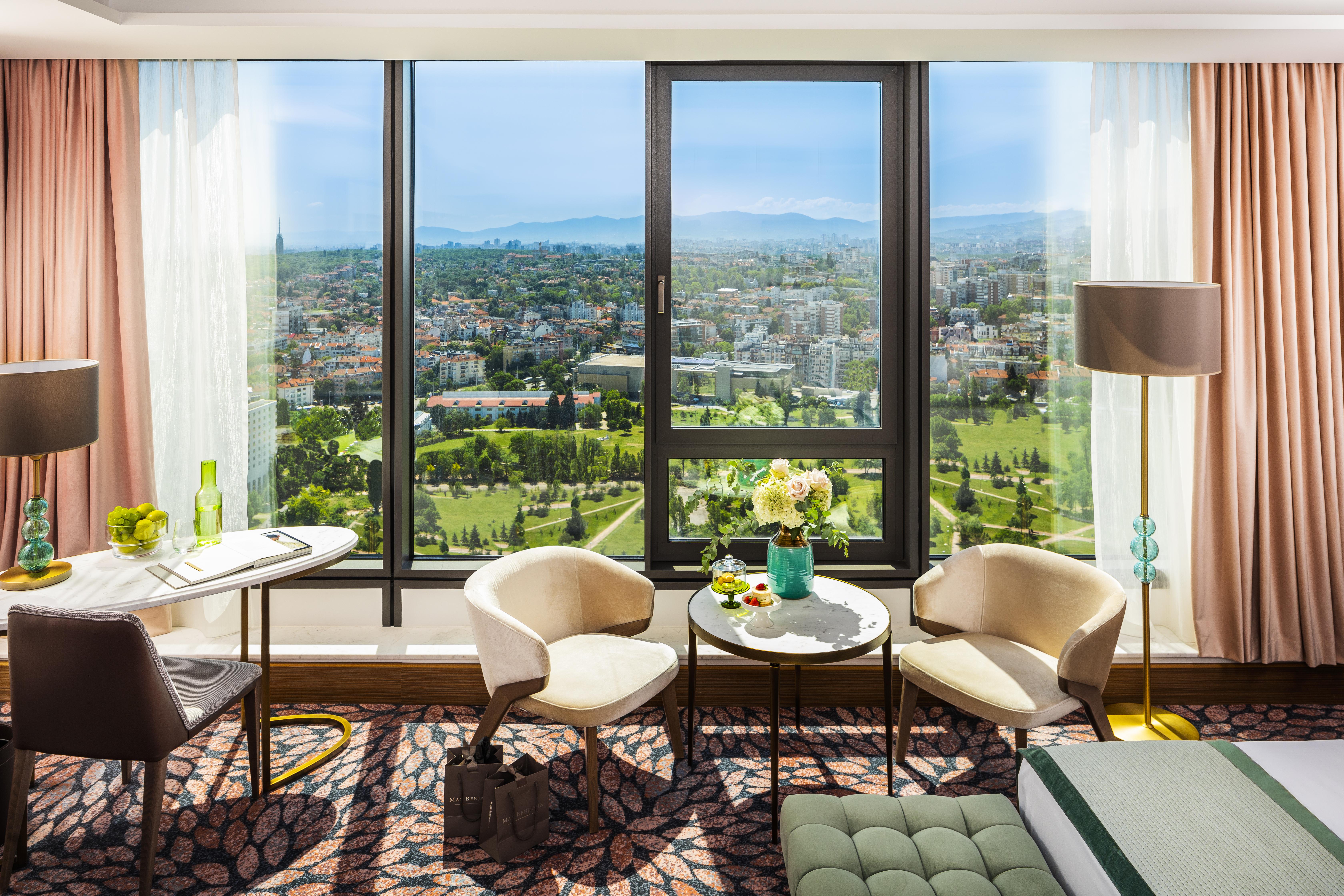 Grand Hotel Millennium Sofia Ngoại thất bức ảnh The photo shows a stylish hotel room with large windows providing a beautiful view of a cityscape and landscape beyond. There are two modern chairs and a small round table set near the windows, accompanied by a vase of fresh flowers. The room feature