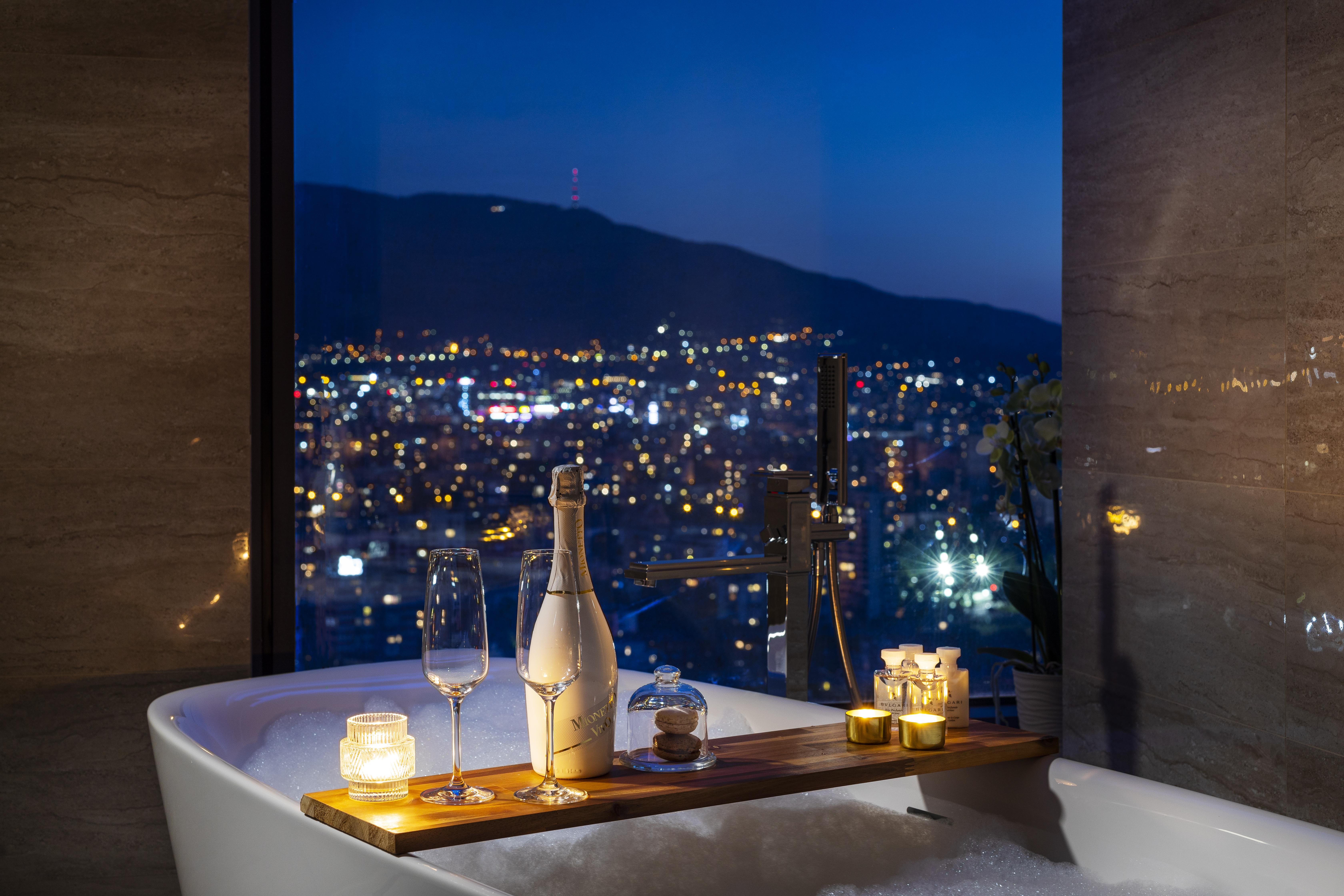 Grand Hotel Millennium Sofia Ngoại thất bức ảnh The photo features a luxurious bathtub placed by a large window, showcasing a scenic city view at night. There is a wooden tray resting on the edge of the tub, displaying a bottle of champagne, two glass flutes, and several lit candles, creating an a