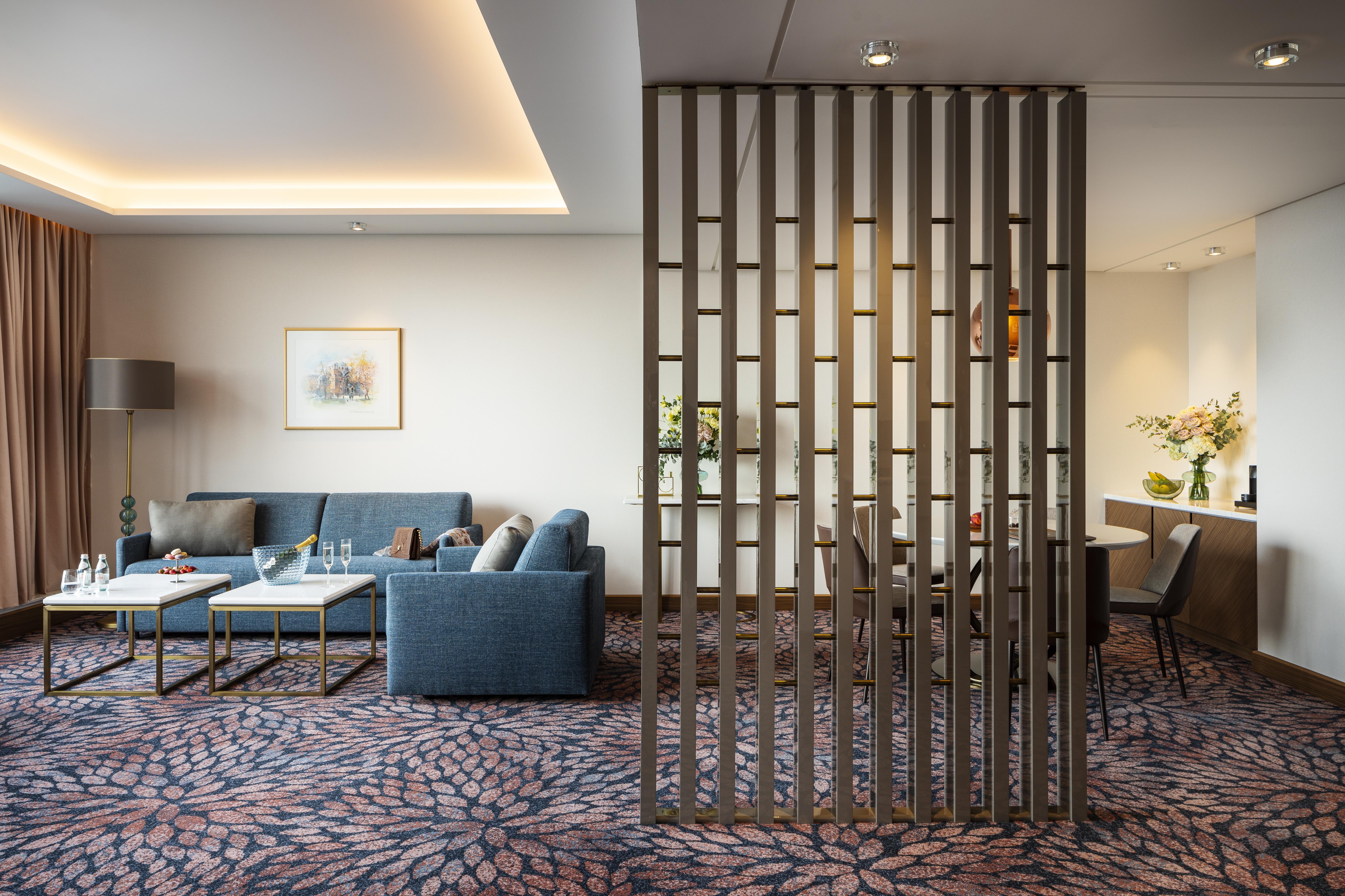 Grand Hotel Millennium Sofia Ngoại thất bức ảnh The photo shows a modern living space with a comfortable seating area featuring a blue couch and two coffee tables. There is a decorative partition made of vertical wooden slats, which separates this sitting area from a dining space. The dining area 