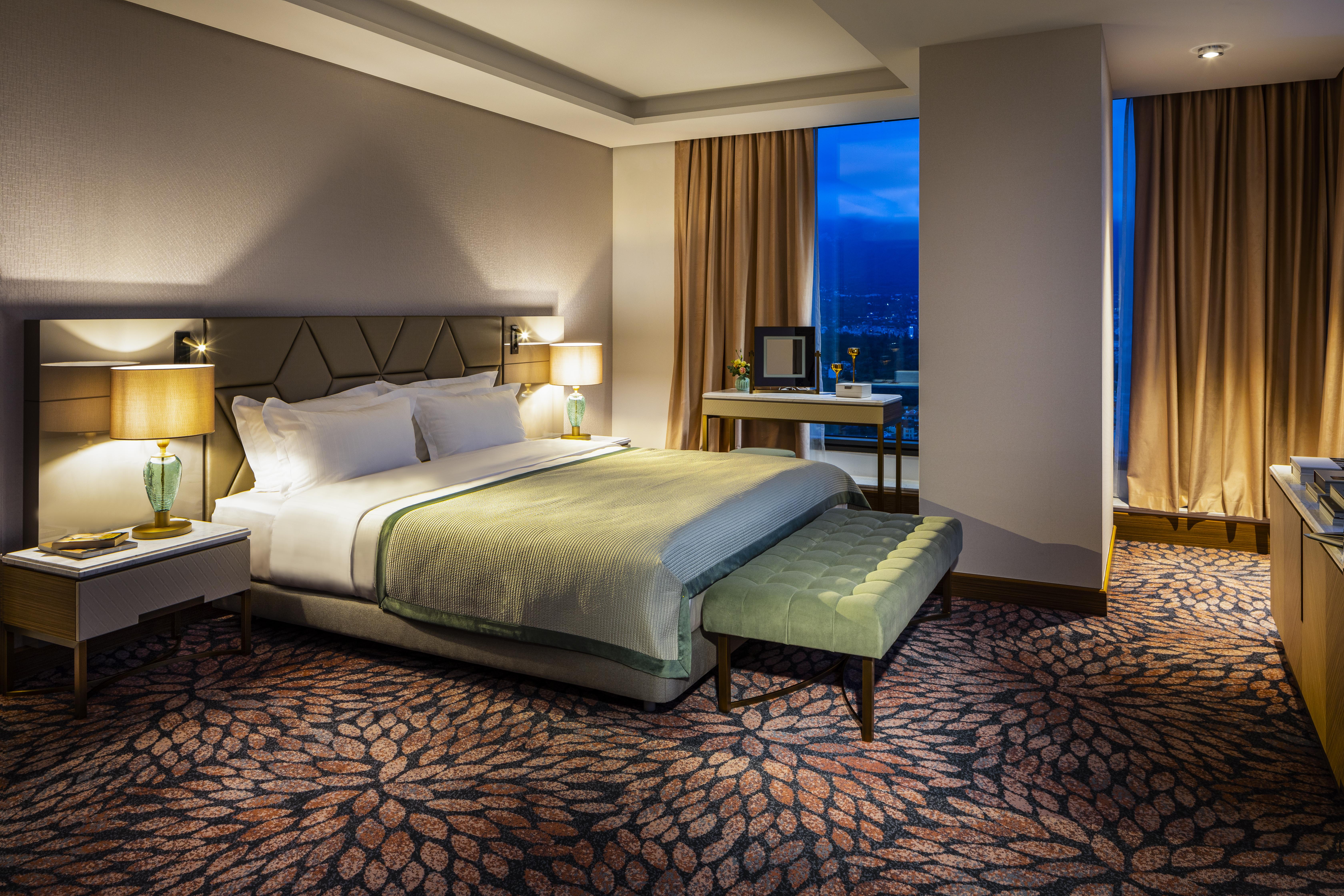 Grand Hotel Millennium Sofia Ngoại thất bức ảnh The photo shows a modern hotel room featuring a spacious design. It has a large bed with a light green throw blanket positioned in the center. On either side of the bed, there are stylish bedside tables with lamps. 

The floor has a patterned carpet,