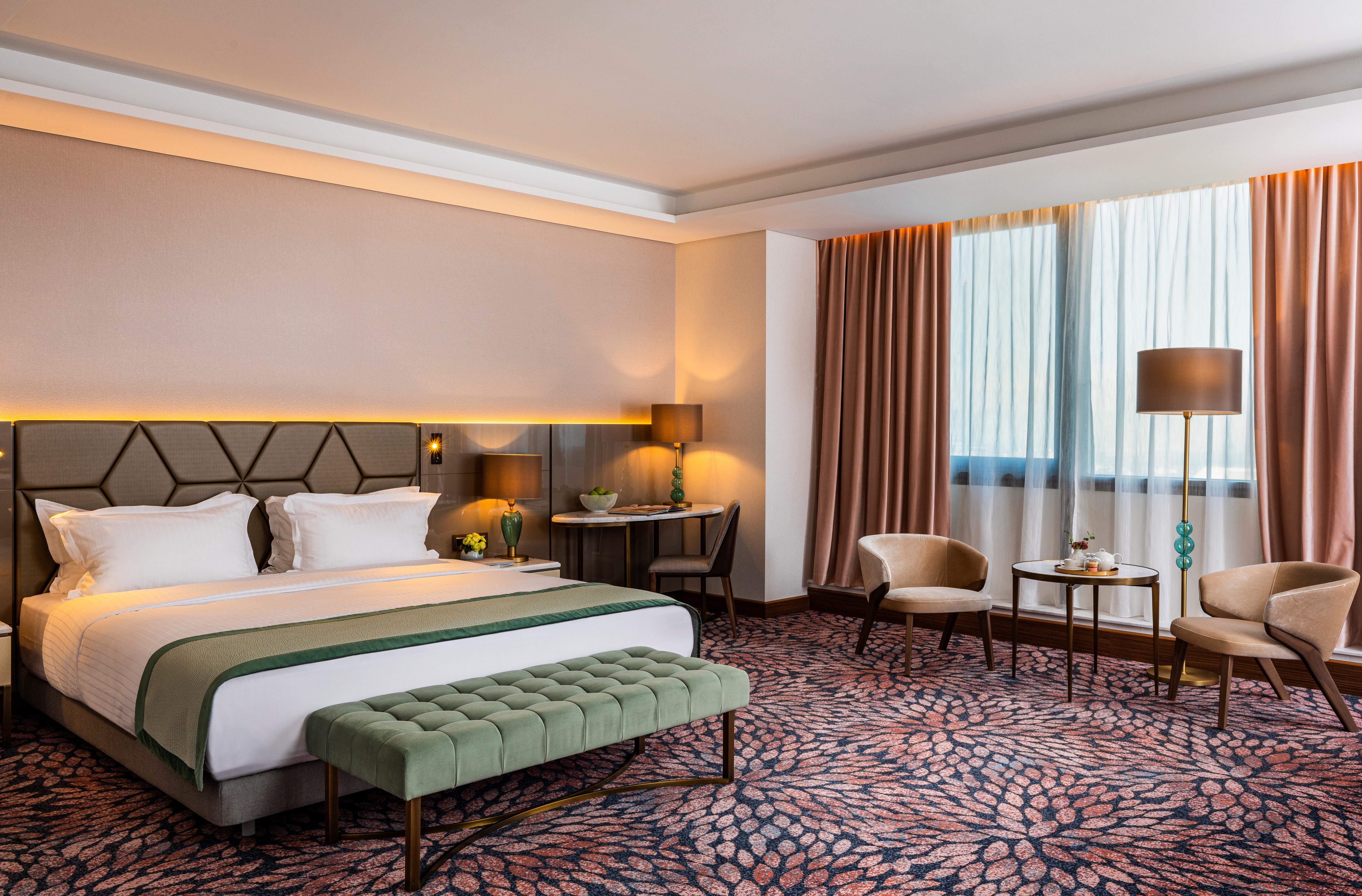 Grand Hotel Millennium Sofia Ngoại thất bức ảnh The photo shows a modern hotel room. It features a large bed with a headboard, topped with two white pillows and a green throw blanket. In front of the bed, there is a stylish green bench. On either side of the bed, there are bedside tables with lamp
