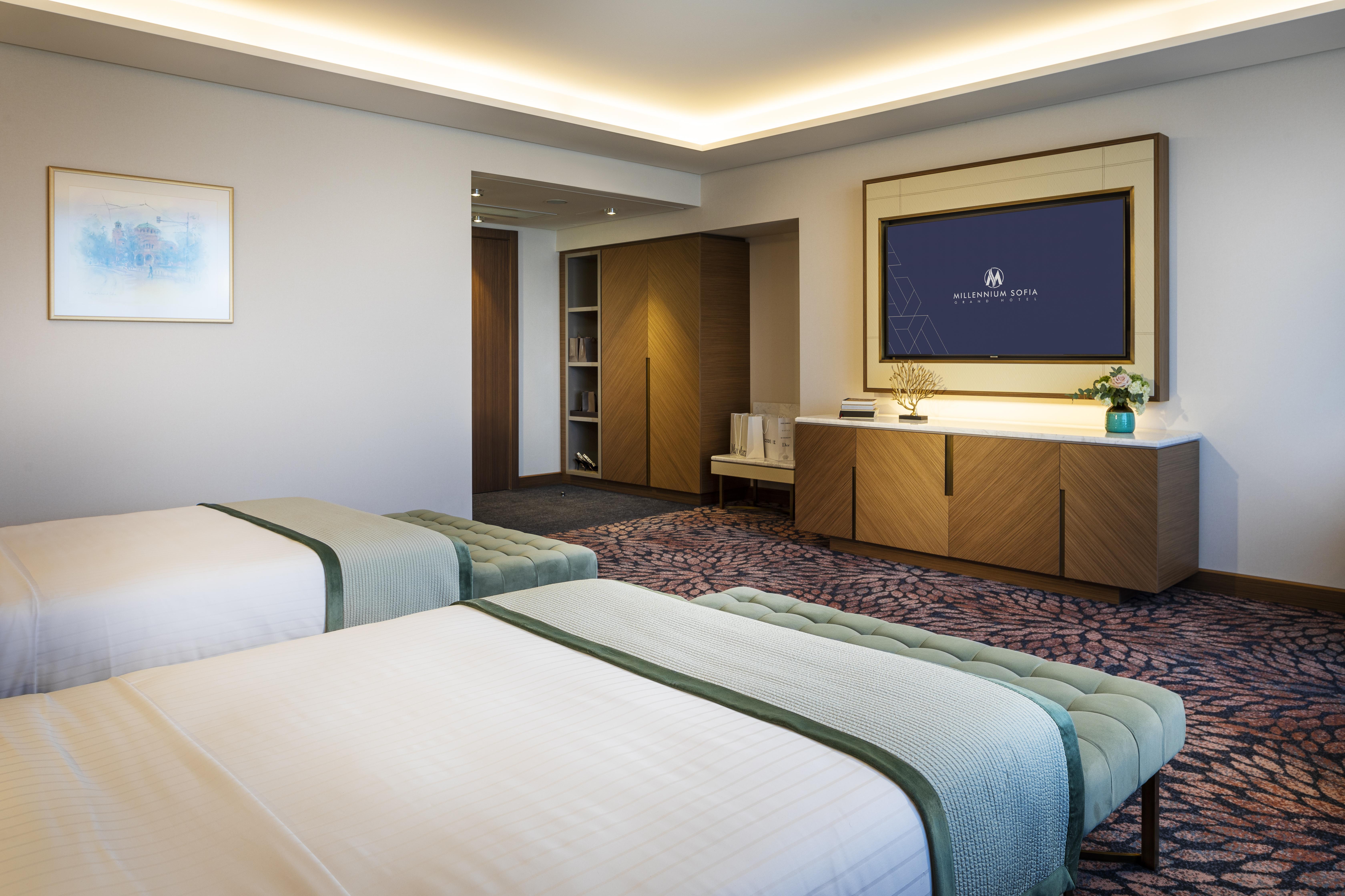Grand Hotel Millennium Sofia Ngoại thất bức ảnh The photo shows a modern hotel room featuring two beds with light-colored bedding and decorative throws. The room has a contemporary design, characterized by neutral walls and a plush carpet. A large television is mounted on the wall across from the 