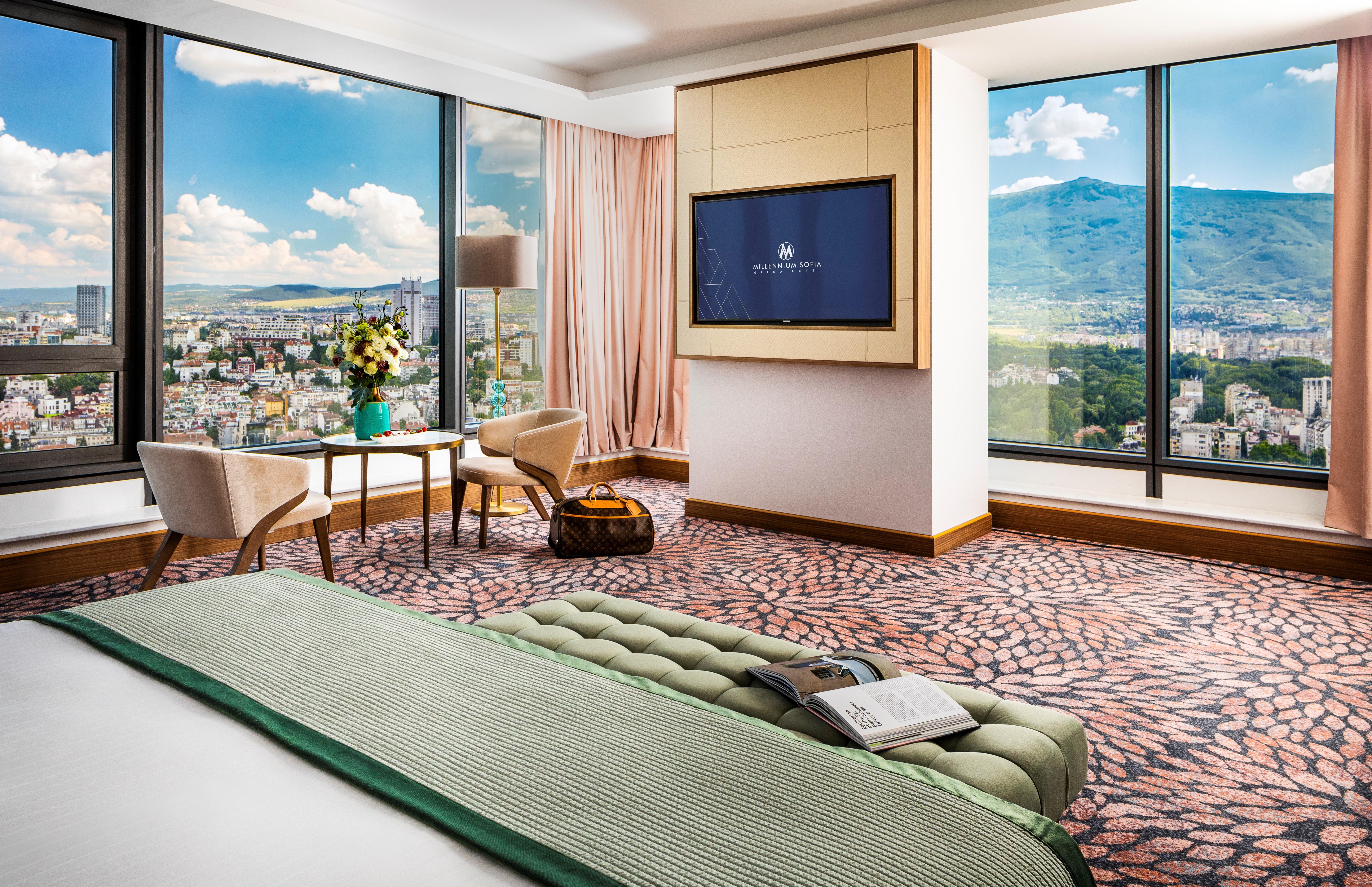 Grand Hotel Millennium Sofia Ngoại thất bức ảnh The photo showcases a stylish hotel room with modern decor. It features large windows providing a panoramic view of a cityscape and mountains in the background. Inside, there is a comfortable bed with a green bedspread, a seating area with two armcha