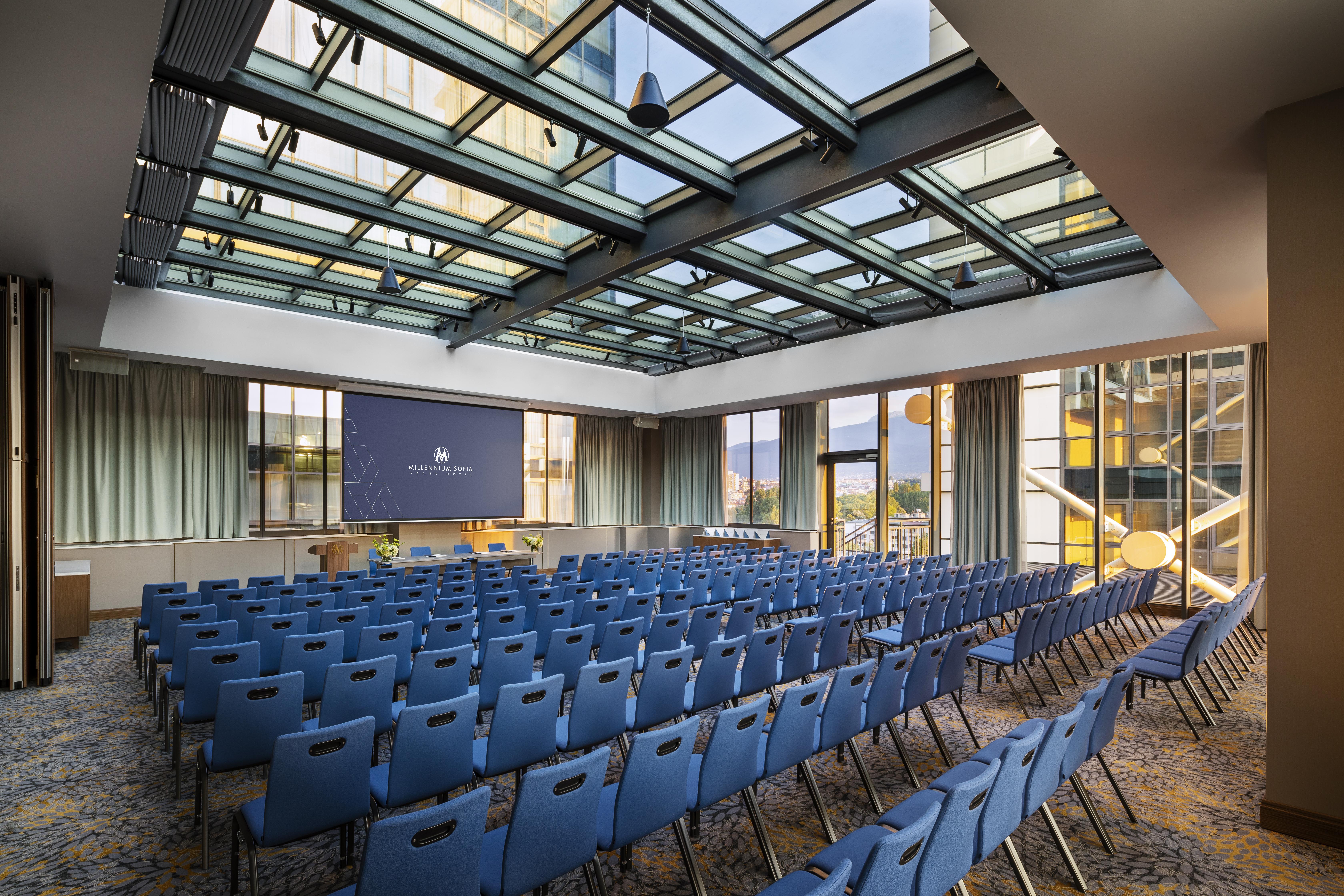 Grand Hotel Millennium Sofia Ngoại thất bức ảnh The photo shows a large conference room or meeting space. It features rows of blue chairs arranged in a theater style, facing a large screen at the front of the room. The room is well-lit, with a glass ceiling allowing natural light to filter in. The