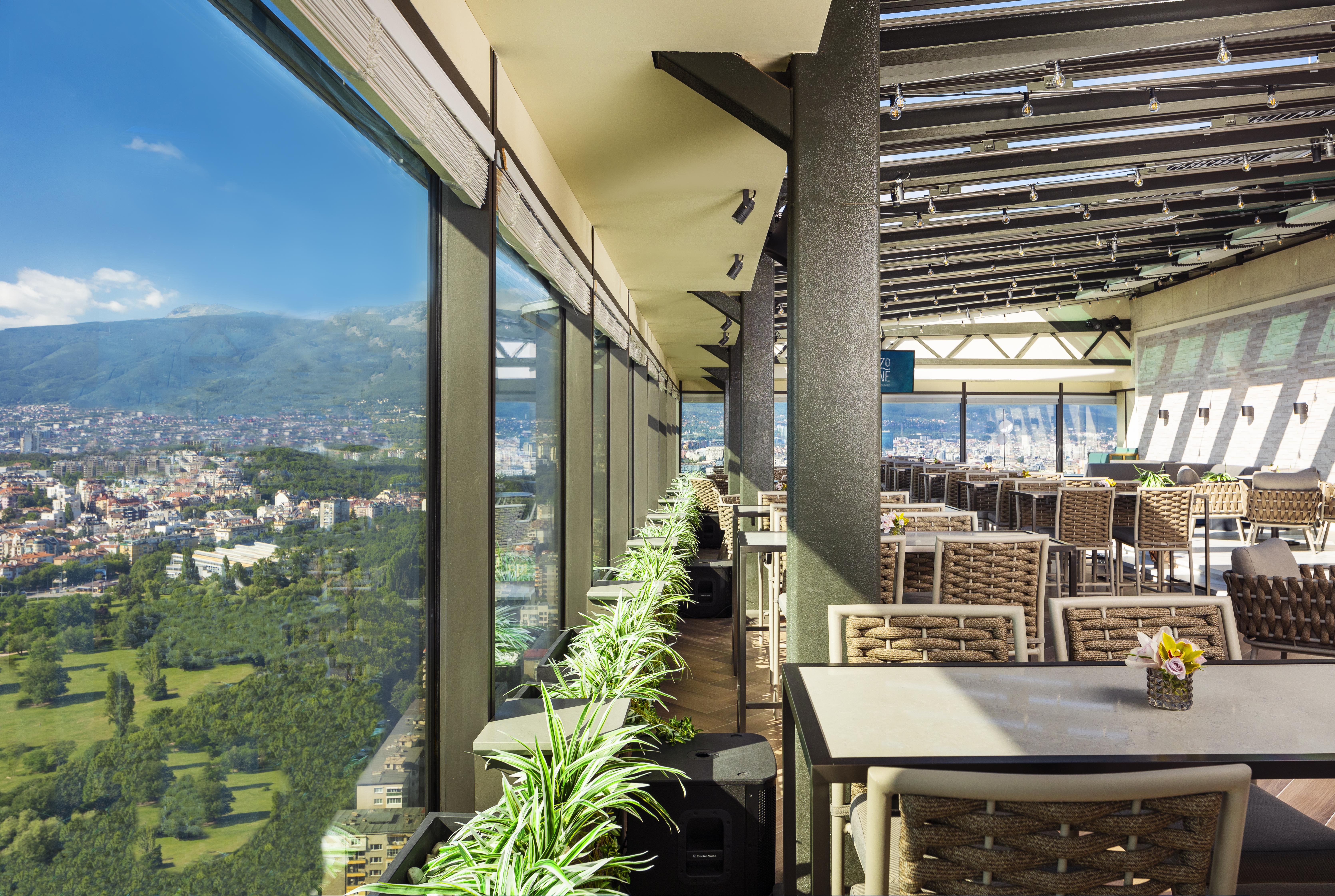 Grand Hotel Millennium Sofia Ngoại thất bức ảnh The photo shows a modern, spacious restaurant with large glass windows offering a panoramic view of a city and surrounding landscape. The interior features contemporary furniture, including stylish tables and chairs. There are potted plants along the